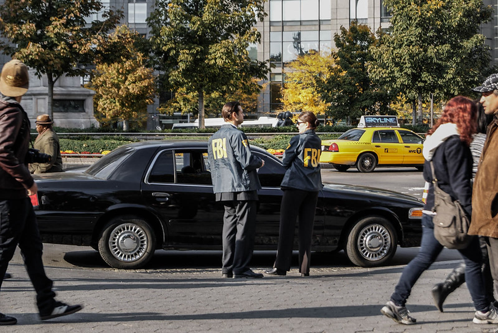 Two people in FBI jackets beside a sedan on a city street.